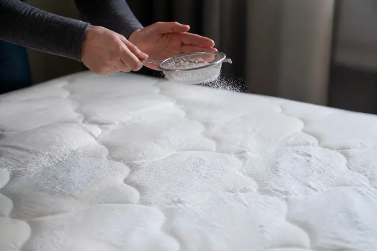 Mains saupoudrant du bicarbonate de soude sur un matelas avec une passoire fine.