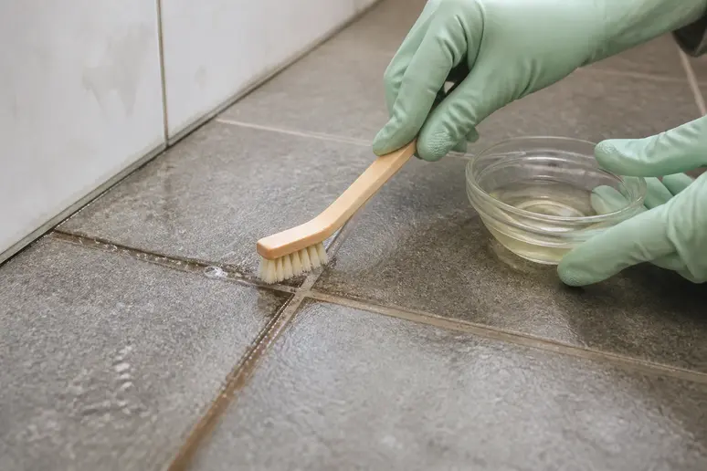 Mains gantées nettoyant un joint de carrelage noirci avec une petite brosse et un produit naturel